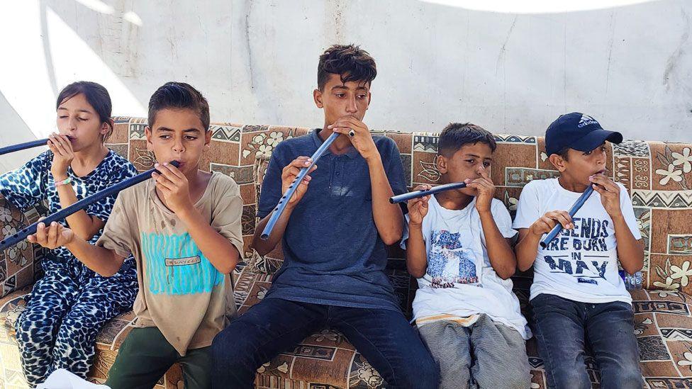 A group of children - four boys and one girl - sit on a brown patterned sofa outside, holding a long straight type of flute to their lips