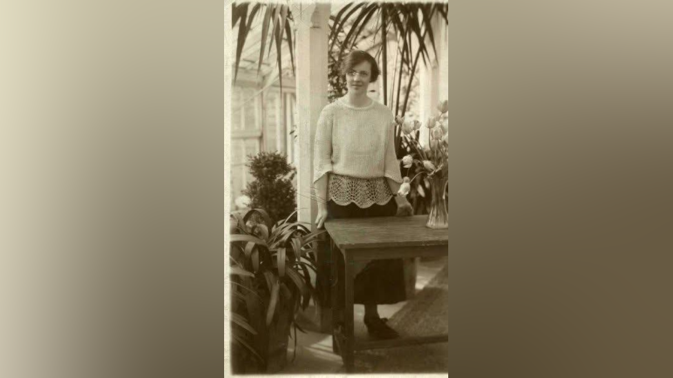 A black and white photo of Mary Schweermann standing in front of a table in the conservatory at Shotley Park. She has short wavy hair and is wearing a white blouse and a black skirt. There are several green plants around her. The tall windows of the room are visible behind her. 