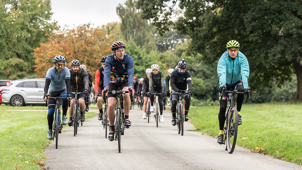 Cyclists riding out of Sandon Hall, flanked by grass and trees