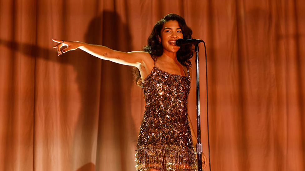 Dean with long, wavy dark hair wearing a sparkly flapper dress, singing into a microphone with one arm stretched out to the side, pointing. Behind her is a gold curtain.