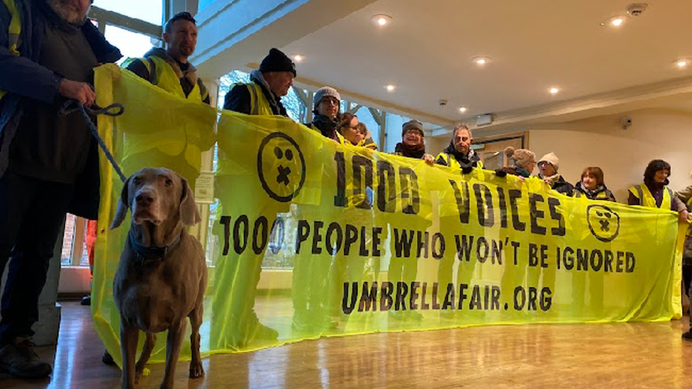 Several people holding a yellow banner saying "1000 people who won't be ignored".  A dog is also present.