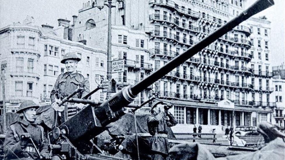 A black and white image of three soldiers and a large anti-aircraft gun in front of a large hotel. 