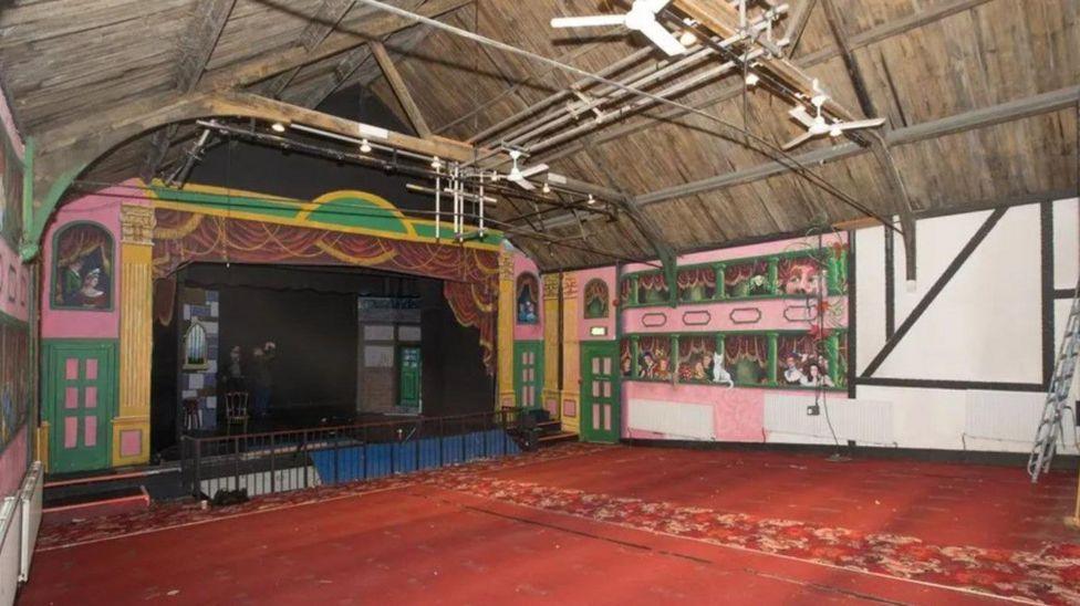 Interior of a theatre with a brick-coloured carpet and a black stage area with a wooden vaulted ceiling and murals on the walls depicting gallery seating full of theatregoers