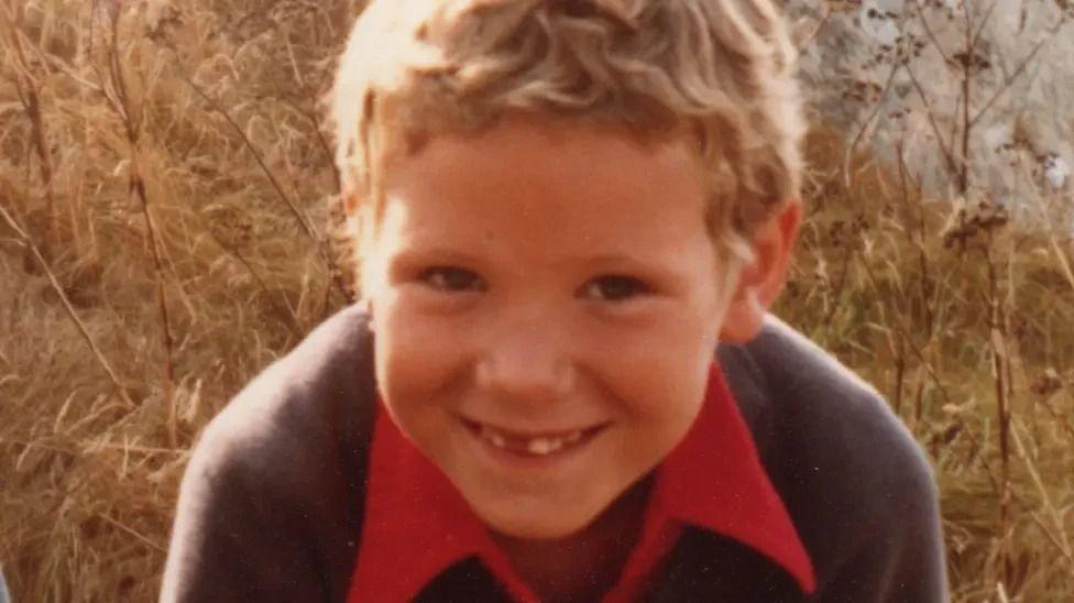 A younger Hamish Cooper smiles for a photo in a field. He is missing some baby teeth and has blond, short wavy hair, wearing a blue jumper over a red polo shirt.