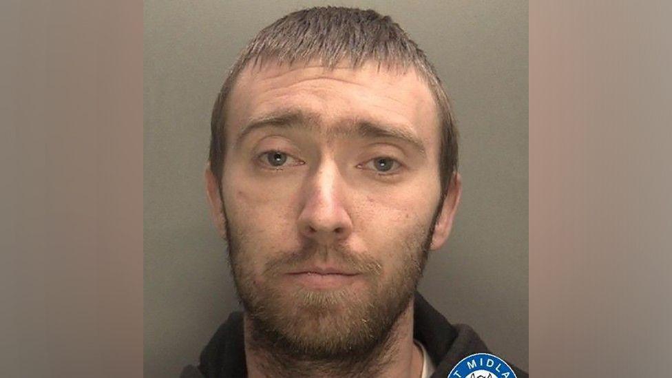 A mugshot of a man with short, brown hair and a beard. He is staring into the camera and wearing a black hoodie.
