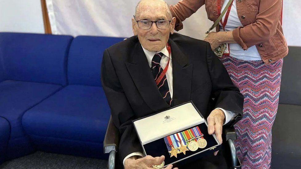 Man wearing glasses and dark coloured suit seated in wheelchair holding a box of medals.