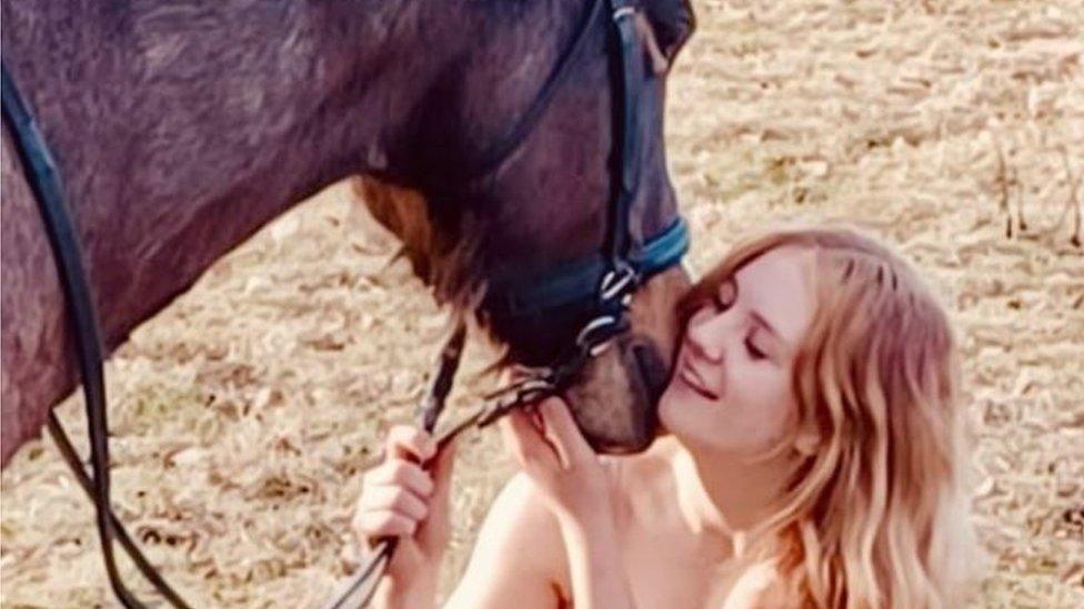 Roxy Phillips nuzzles a large brown horse while holding its reins in her right hand. She has long blonde wavy hair. She is smiling and has her eyes closed as she puts her cheek to the horse's nose and tickles its chin with her other hand.