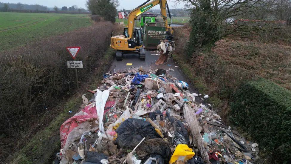 A huge pile of rubbish blocking the road in a rural area of Staffordshire in the UK. A yellow digger is clearing it away, by scooping it into the back of a green lorry.