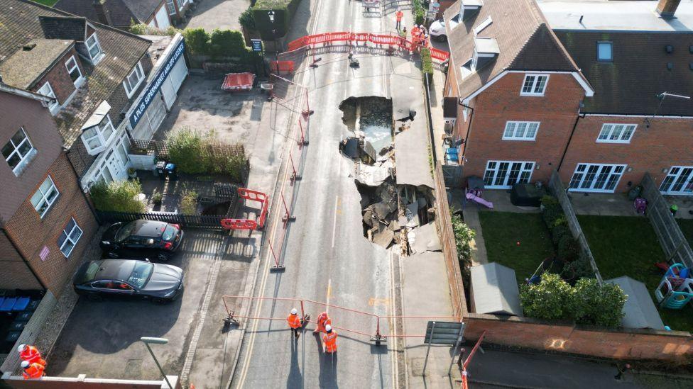 An areal view of the sinkhole. There are buildings either side of the road and a group of workers in high-visibility jackets. 