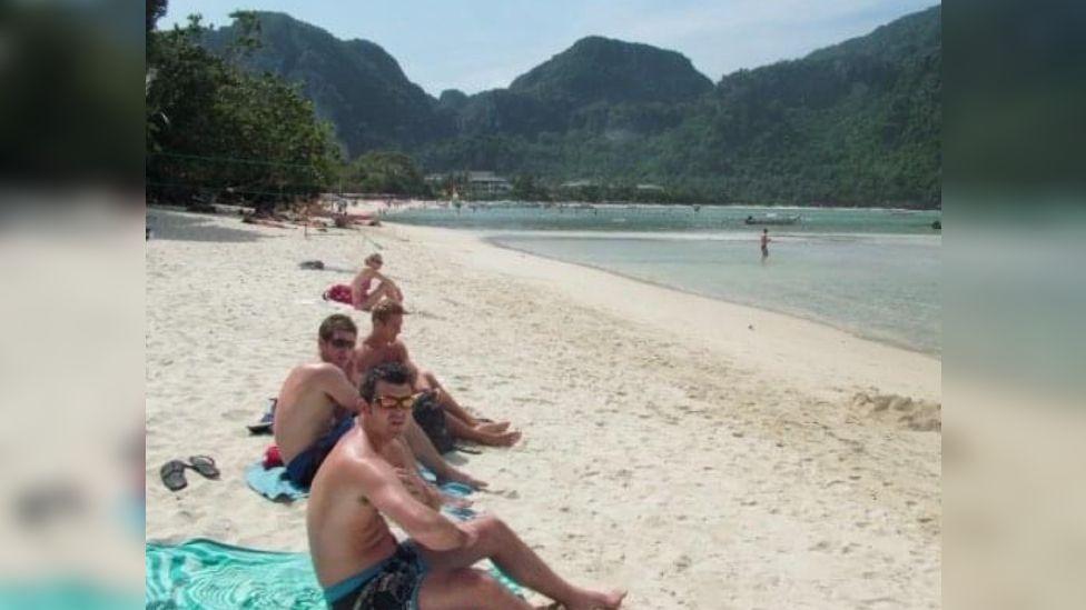 A group of friends sitting on blue towels on the beach. The white sandy cove is tucked between a large green mountain range and the water is very shallow and clear. 