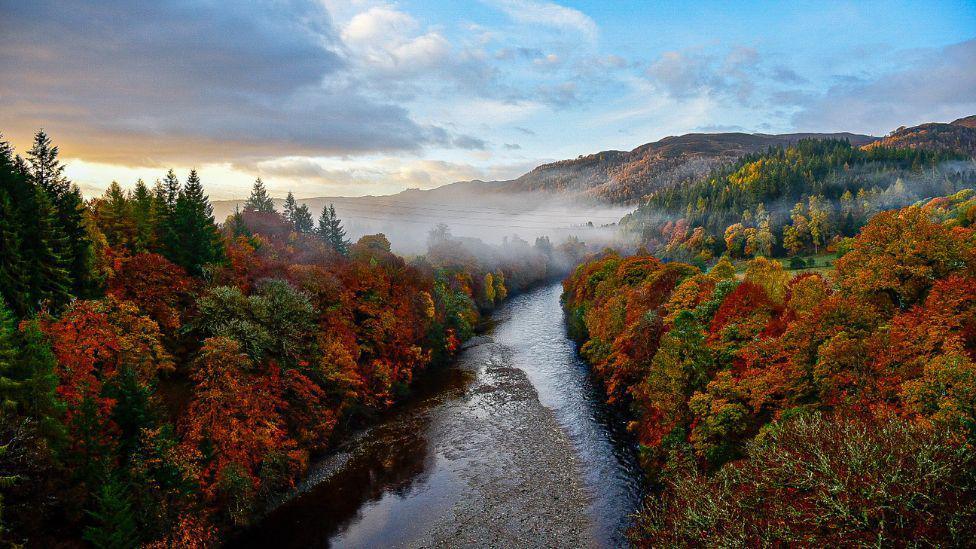 An autumnal scene from Killiecrankie in Perthshire. A river runs through the middle of the scene. On either side, trees coloured orange, yellow, red and green are under a blue sky. There is cloud above the trees on the left side with the light from the sunrise just poking through. In the distance, a layer of fog covers part of the trees and river in front of several hills.