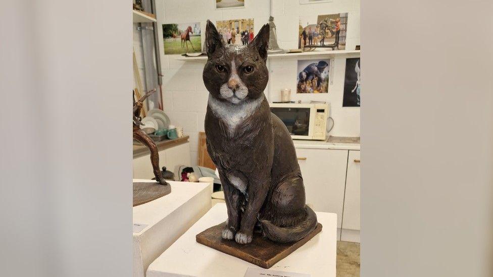 A life-size statue of a black and white cat sitting on a metal plinth. The statue is in an art studio and there are photos of other statues on the wall behind it.