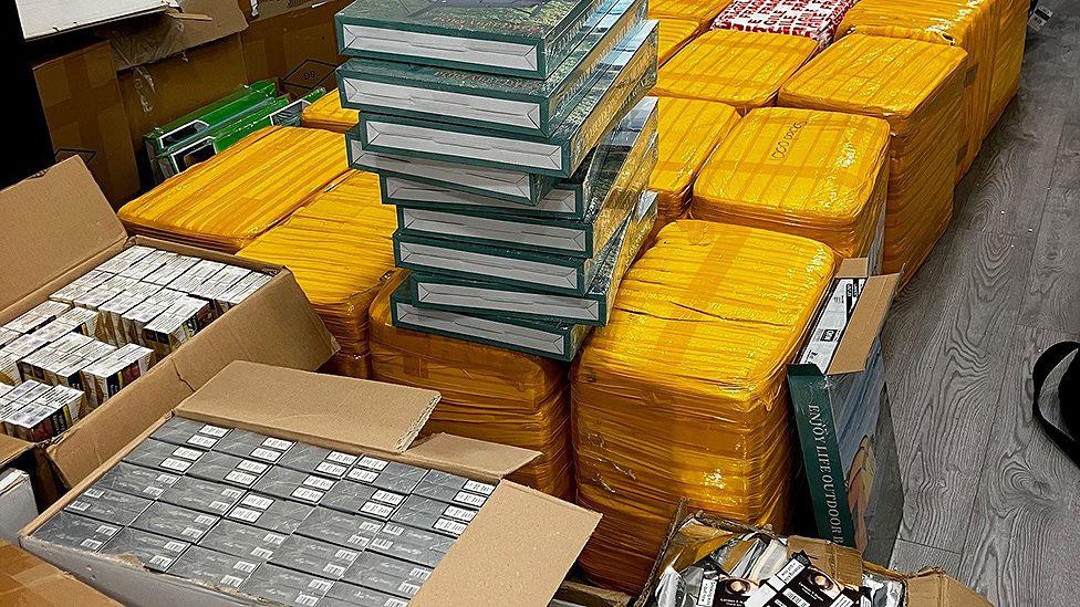 Boxes of cigarettes in front of four rows of large packages on the floor, they are wrapped in yellow tape and there are some green boxes in a pile on top of the first row