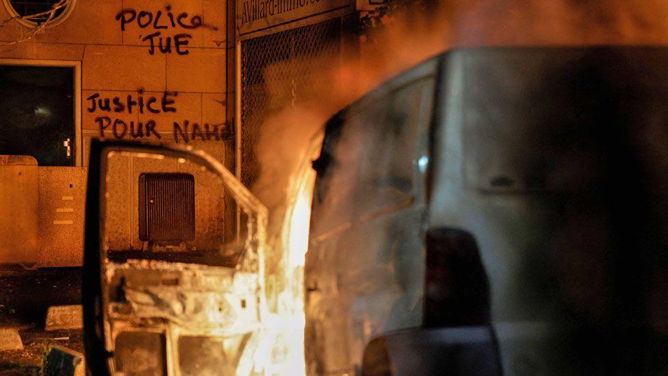 A van burns in front of a building with graffiti reading in French 'Police kill' and 'Justice for Nahel' in Nanterre, France, a day after the killing of 17-year-old Nahel Merzouk - 28 June 2023