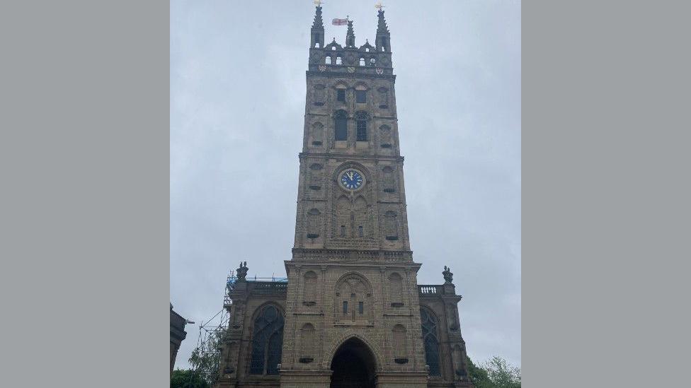 The front entrance of a church. It is made from brown brick and has a blue clock on the front. The English flag is flying on the top