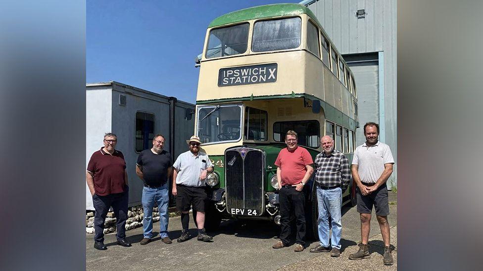 Six gentlemen stand in front of a cream and green double-decker bus smiling. Its licence plate says EPV 24 and the destination sign reads 'Ipswich Station'