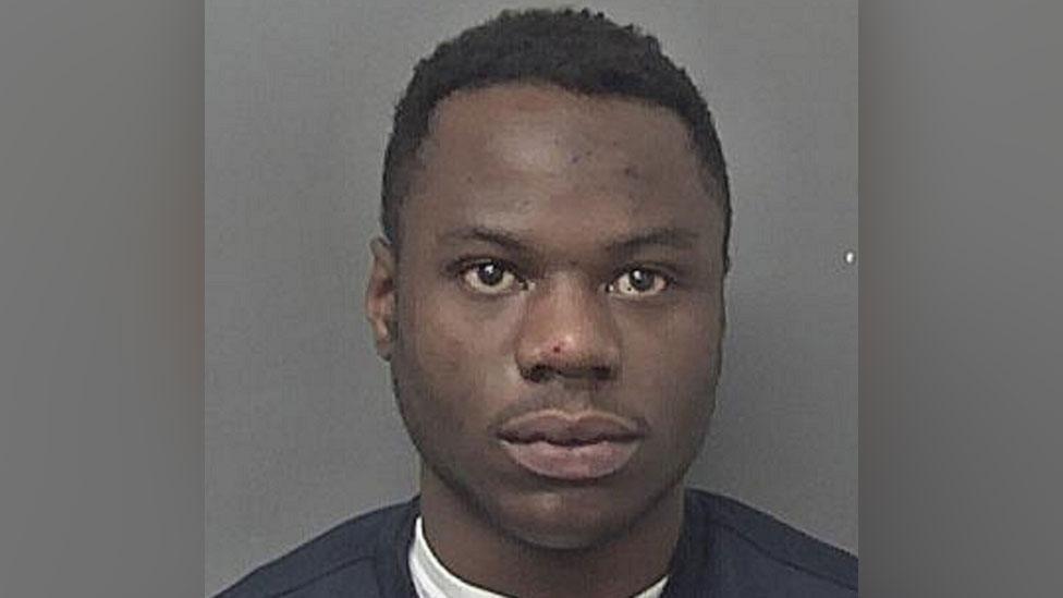 A man wearing a black jumper stands against a grey coloured wall and stares expressionless into the camera.