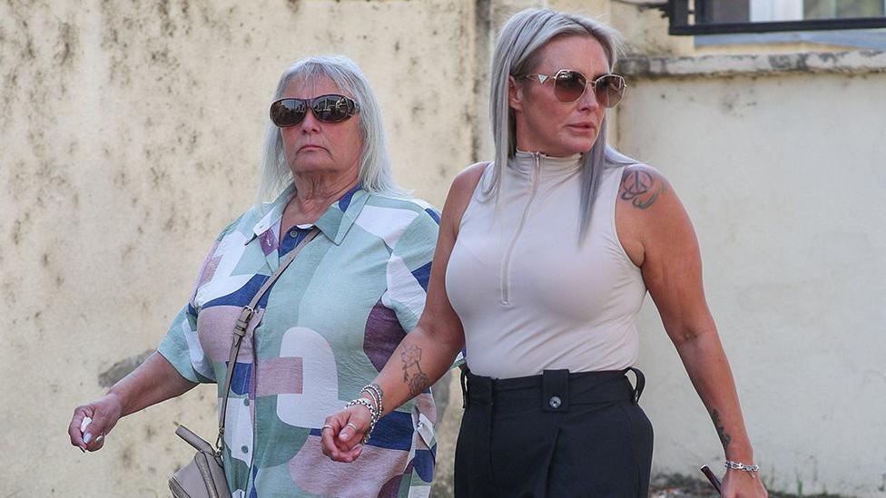 Bella Culley's mother, Lyanne Kennedy, and, on her right, Bella's grandmother, Christine Cook, walking outside the Tbilisi City Court building in Tbilisi in Georgia. Lyanne Kennedy has long, dyed platinum-grey-blonde hair with dark roots showing and is wearing silver necklaces, bracelets and rings, and has on sunglasses. She is wearing smart, black trousers and a pale, pinky-grey sleeveless top with a front zip, zipped up, and has tattoos on her left shoulder and right forearm. Christine Cook is wearing a blue, green, white and purple geometric design shirt and has shoulder length, wispy, white hair. She is wearing dark sunglasses and has a stern expression.