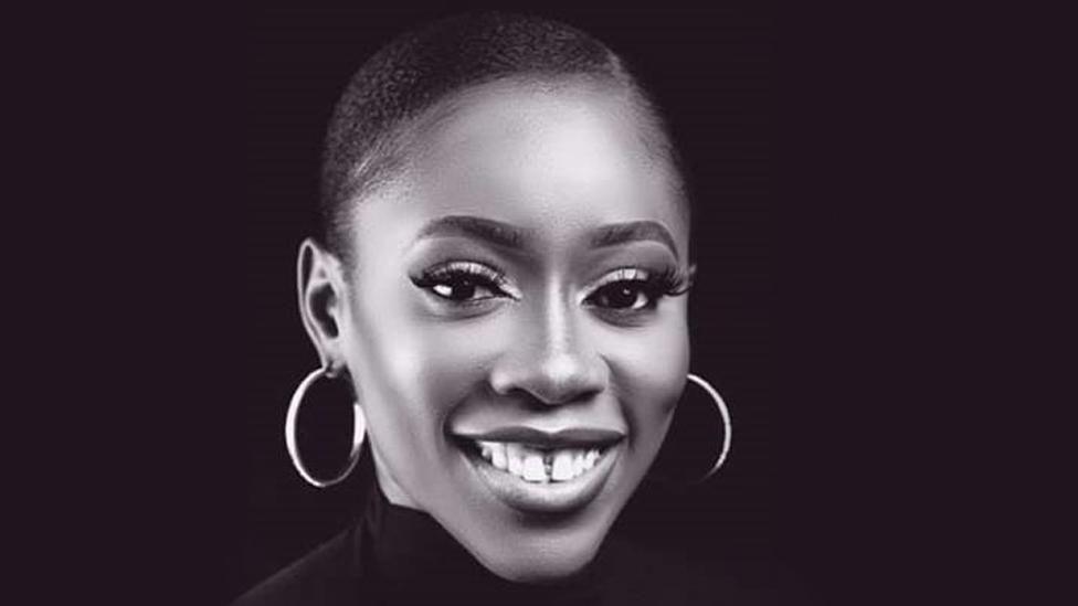 Chinenye Okonkwo is smiling at the camera. Her black hair is scraped back and she is pictured against a black backdrop. She is wearing large, hooped earrings