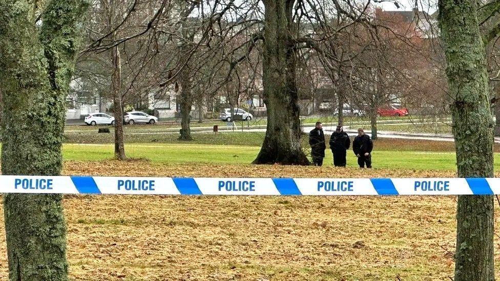 Blue and white police tape between trees in a park, with police officers in the background.