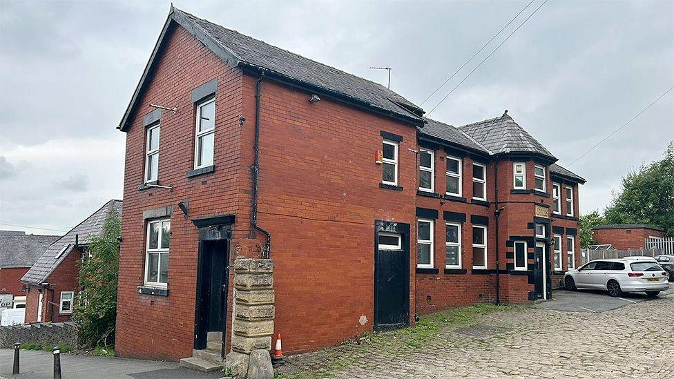 Street view image of the Masjid Al Aqsa mosque Blackburn, a red brick two-storey building with white windows and a cobbled side alley