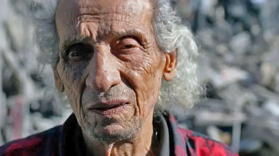 A man, Fouad Hassan, stares off into the distance. Behind him is a twisted pile of concrete and metal rubble. Some of his family used to live where the rubble is now piled high.