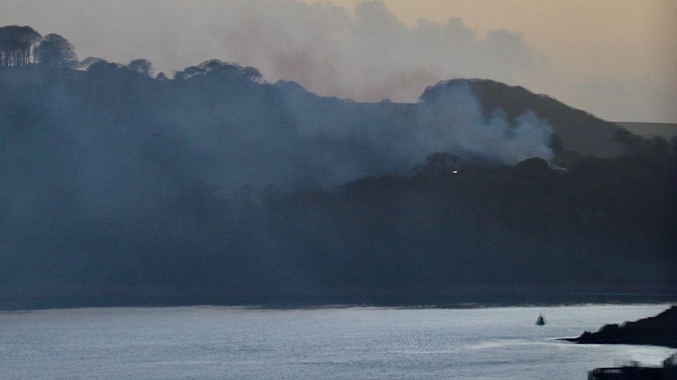 Smoke is rising from a hill seen from across a river