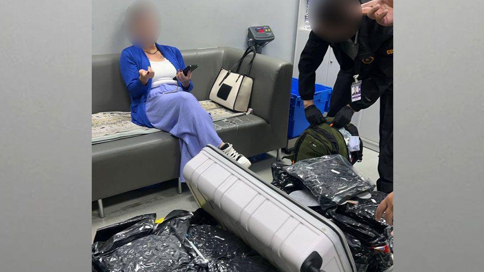 A woman sits on a sofa gesturing with one hand whilst holding a phone in another, her legs crossed, and her face is blurred. Meanwhile some in uniform is searching through a bag full of black vacuum compressed bags.