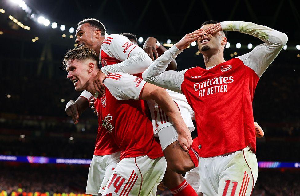 A group of Arsenal players in red and white jerseys celebrating a goal on the pitch, with one player saluting and others embracing under stadium lights.