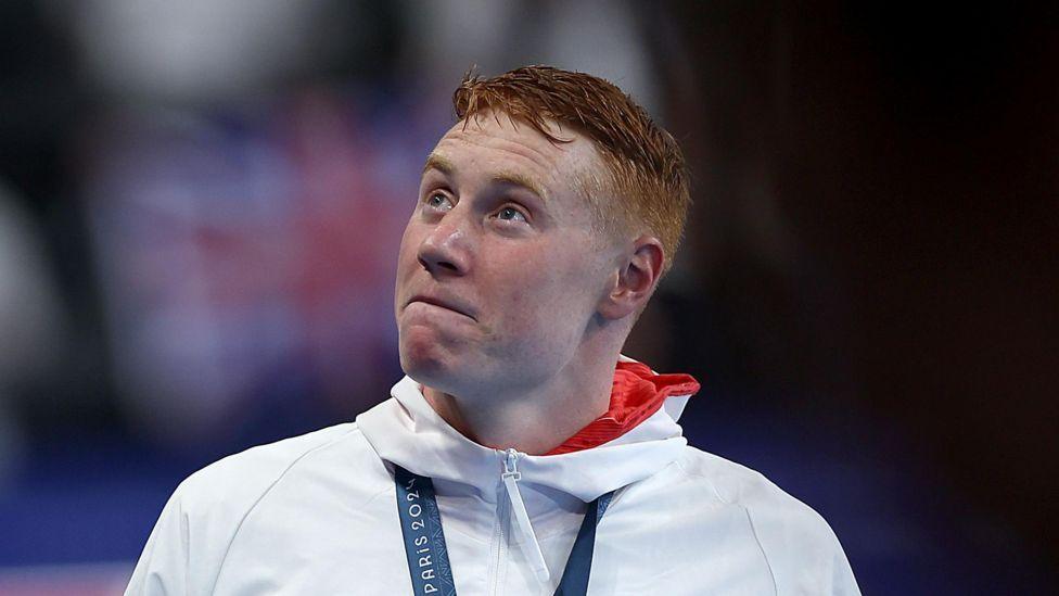 Tom Dean, who has red hair and wears a white and red hoodie with a medal around his neck, looks to the side, with a Union Flag seen in the background 