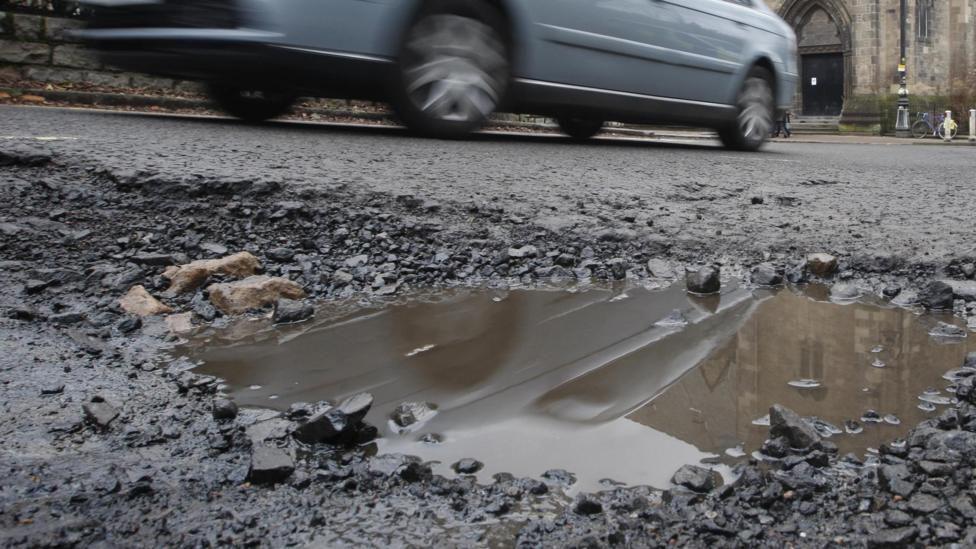 A ground-level image of a pothole in a road, full of water. A light blue car is driving past in the background. The entrance to a building is also visible in the distance.