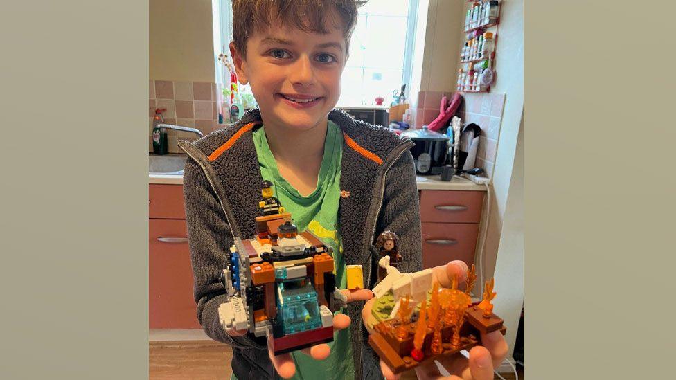 A primary school aged boy Noah standing in a kichen with short mid-brown hair, wearing a fleece and smiling at the camera. He is holding two Lego models, one showing a futuristic fire engine design, the other showing flames.