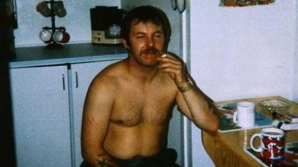 Andrew Walker is sitting at a table which has two mugs on it and an ash tray. He is topless and is smoking. He has brown hair and a brown moustache.