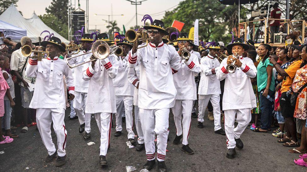 A group of men dressed in white outfits and black large brimmed hats blow trumpets as they parade down a street in Calabar, Nigeria