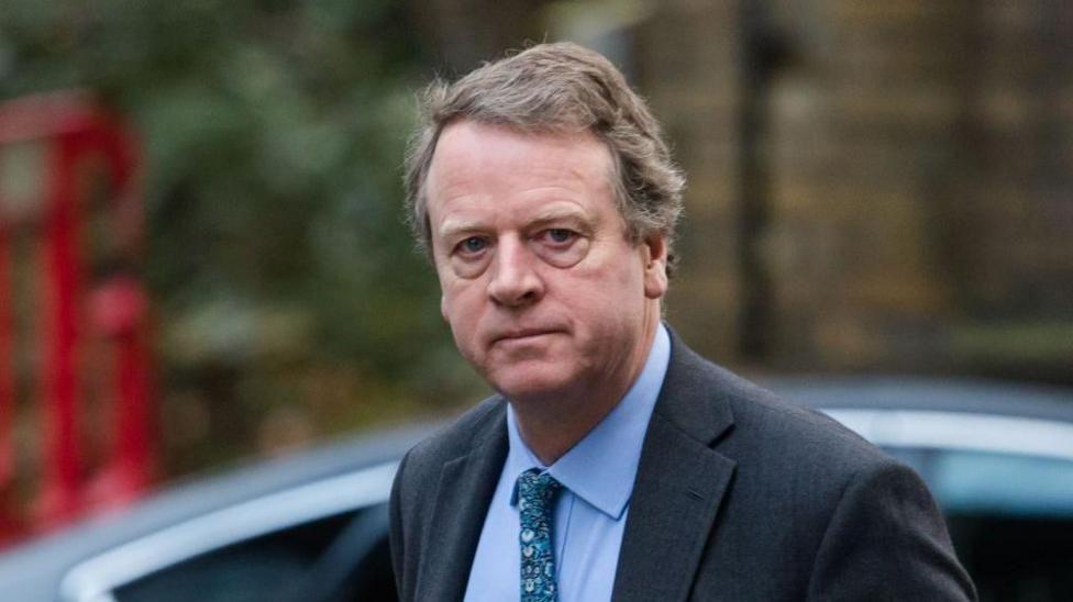 Alister Jack - a man with greying fair hair, wearing a dark suit with a blue shirt and patterned tie - pictured shortly after getting out of a car in London