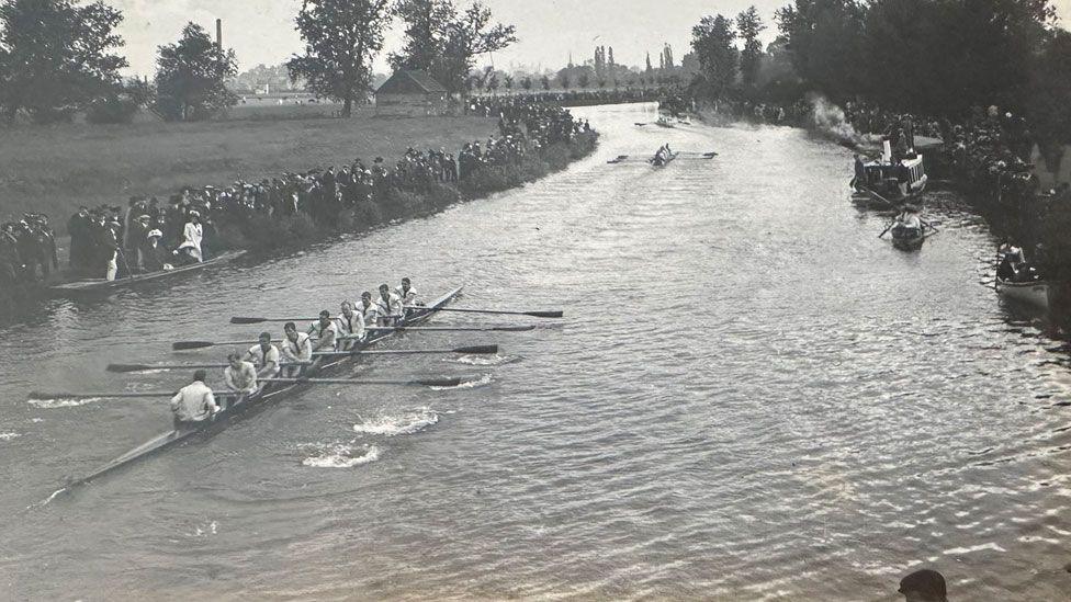 A black and white image showing rowers competing on the River Cam. Crowds of people are gathered on the river side. Behind them can be seen grass, trees and the tall chimney of the sewage pumping station. The river bends around the corner.