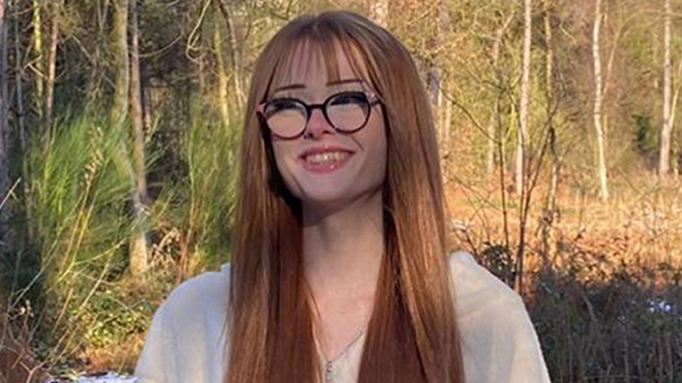 Brianna Ghey, with long red hair and black glasses, standing in a forest with trees around her