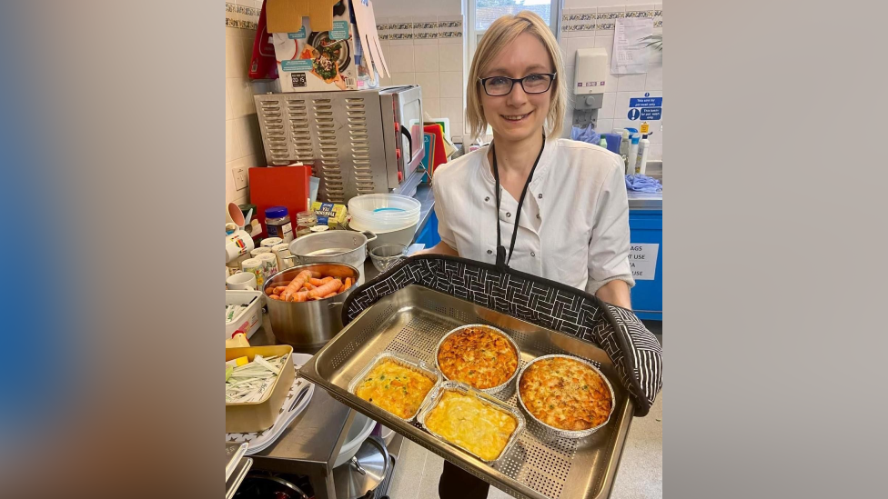 Hannah Hobbs-Chell, who has blonde hair, black-rimmed glasses and a white shirt, is standing and smiling while holding a metal tray with oven gloves.