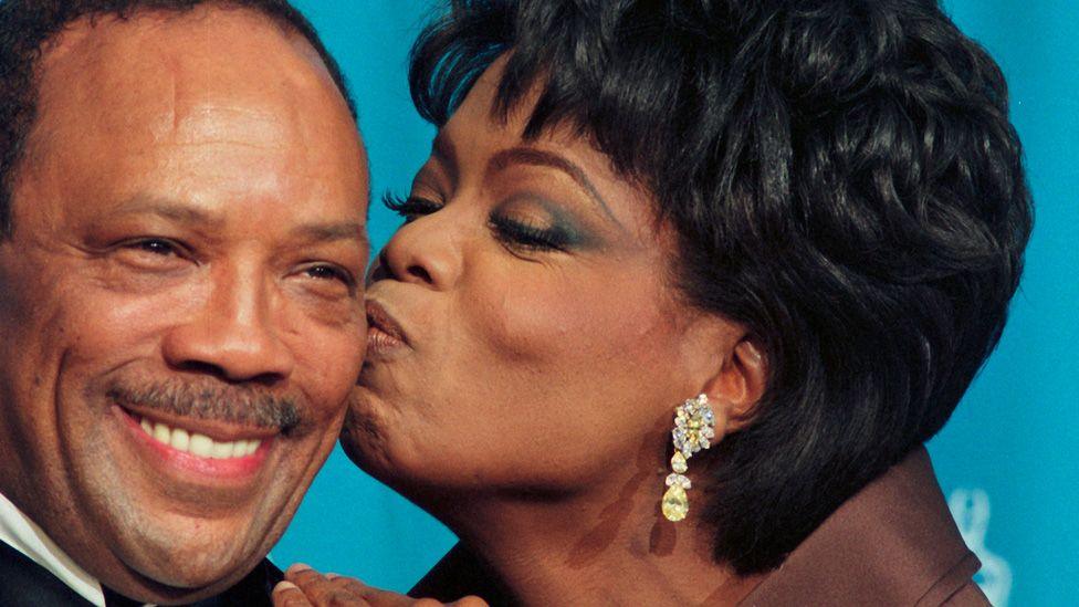 Oprah Winfrey kissing a smiling Quincy Jones on the cheek backstage at the 1995 Oscars 