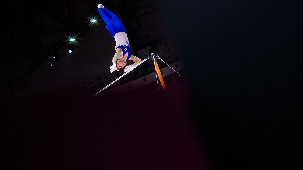 Gymnast in a blue and white uniform performs a high bar routine, captured mid-flip above the apparatus under bright spotlights in an indoor arena.