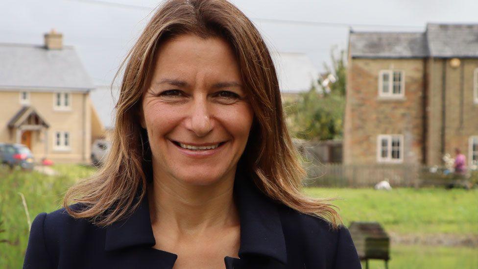 Lucy Frazer wearing a blue jacket looking towards the camera with houses and a village green behind her