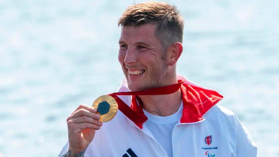 Gregg Stevenson holding up a gold medal and smiling in a white tracksuit