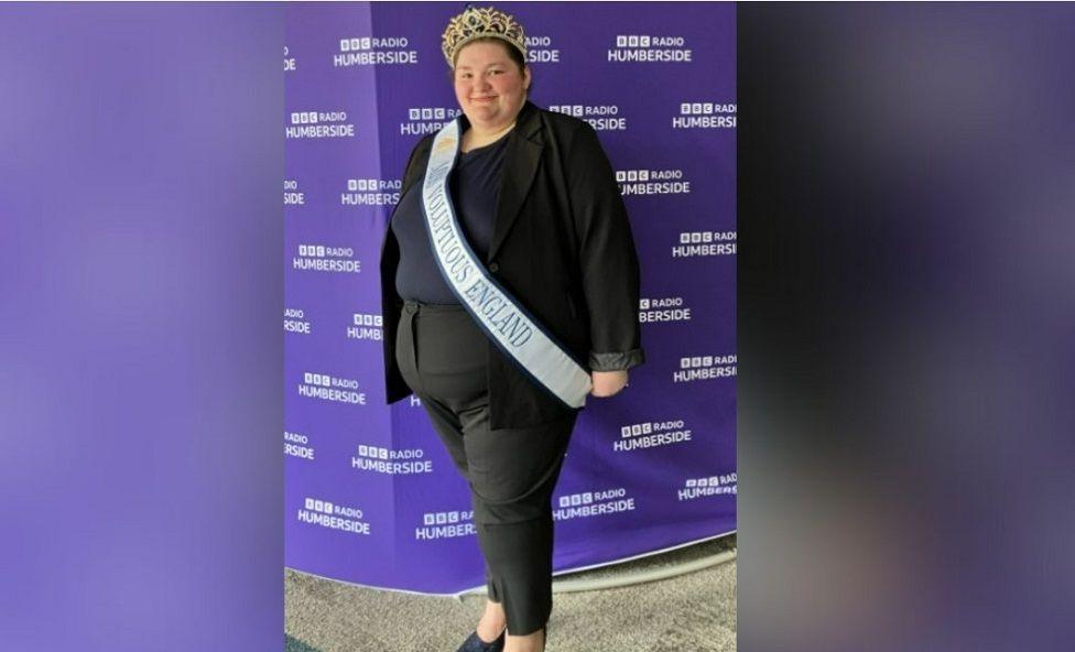 A full-length image of Philippa Mackinder, showing more of her sash and her outfit, which includes a black blazer over a navy top and black trousers and shoes. Her sash reads "Miss Voluptuous England". She is wearing a gold crown on her head. She is standing in front of a purple BBC Radio Humberside backdrop.