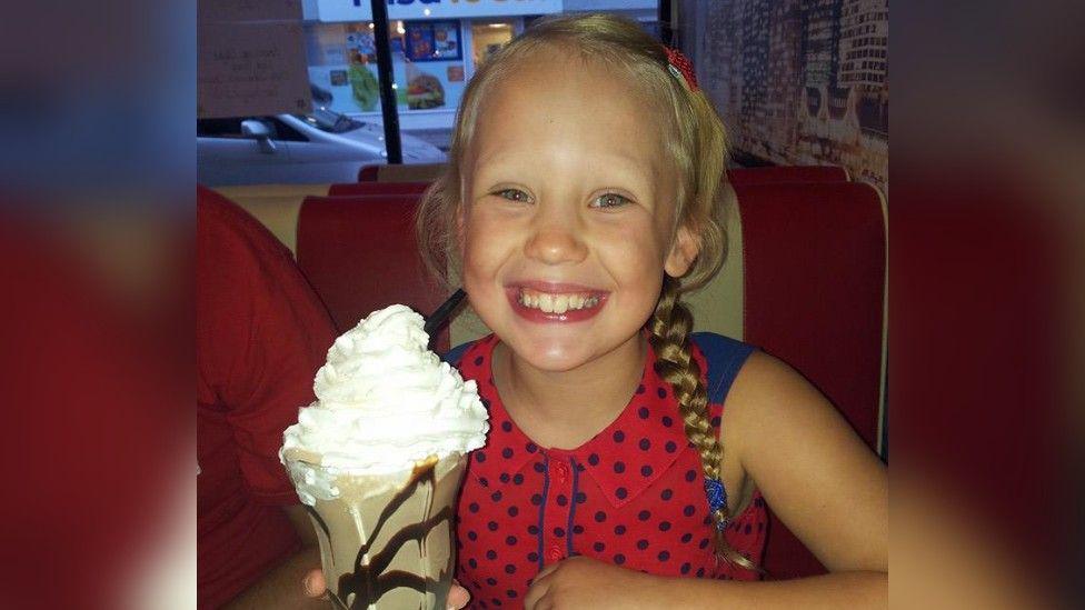 A young girl with a wide teethy smile sat in a red diner chair in a cafe holding a large ice-cream sundae. She has blonde hair in plaits and a red and blue polka-dot top on.