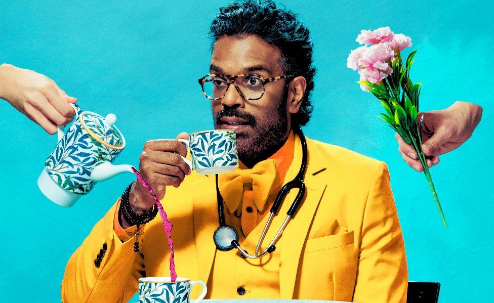 Romesh Ranganathan pictured in promotional image for Woman in Mind, wearing a bright yellow suit with a stethoscope around his neck, drinking a cup of tea