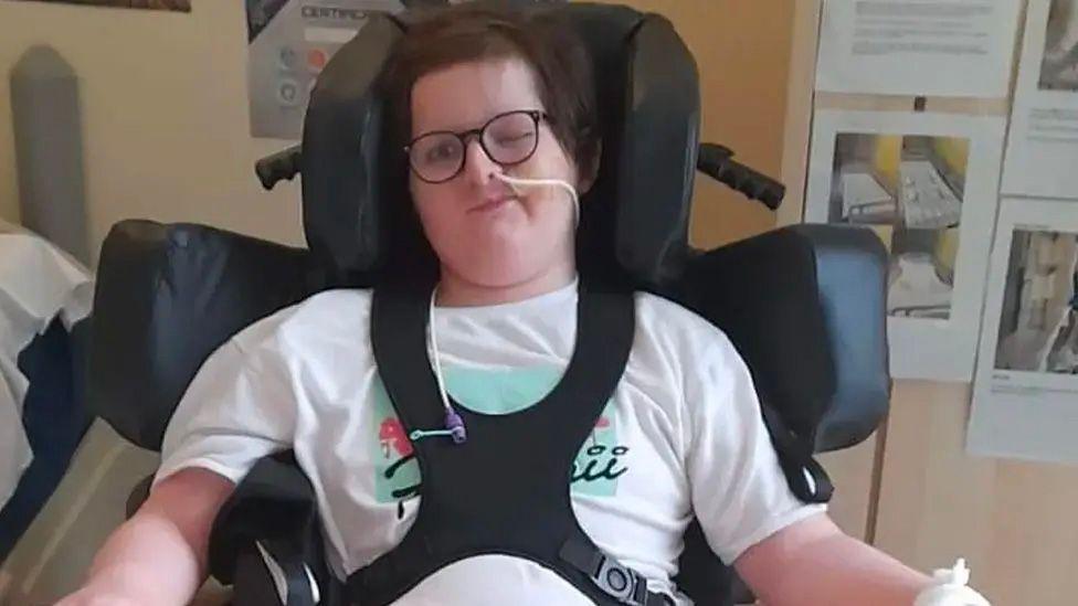 Daniel Flemen in a hospital room strapped into a wheelchair with head and shoulder supports. He has dark hair and glasses and a tube in his nose.