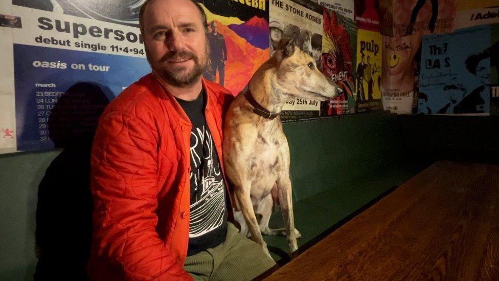 Paul is wearing a black t-shirt showing an artistic illustration of a ship under a red jacket and is holding a lurcher dog, which is looking away to the right