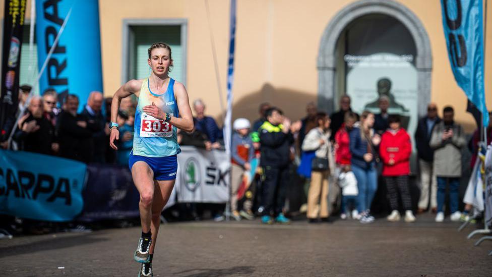 A runner strides out in racing vest and shorts with a crowd of spectators lined up in the background