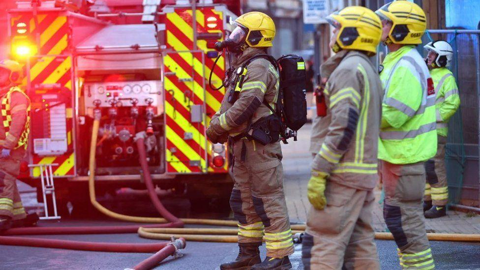 Four firefighters stand to the side of a fire engine with hoses snaking on the road