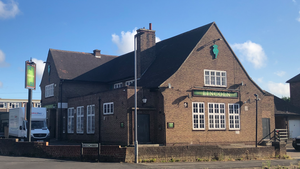 The exterior of the Lincoln Imp pub in Scunthorpe. The dark brick building is bathed in sunshine with blue skies. A van is parked next to a large green sign.